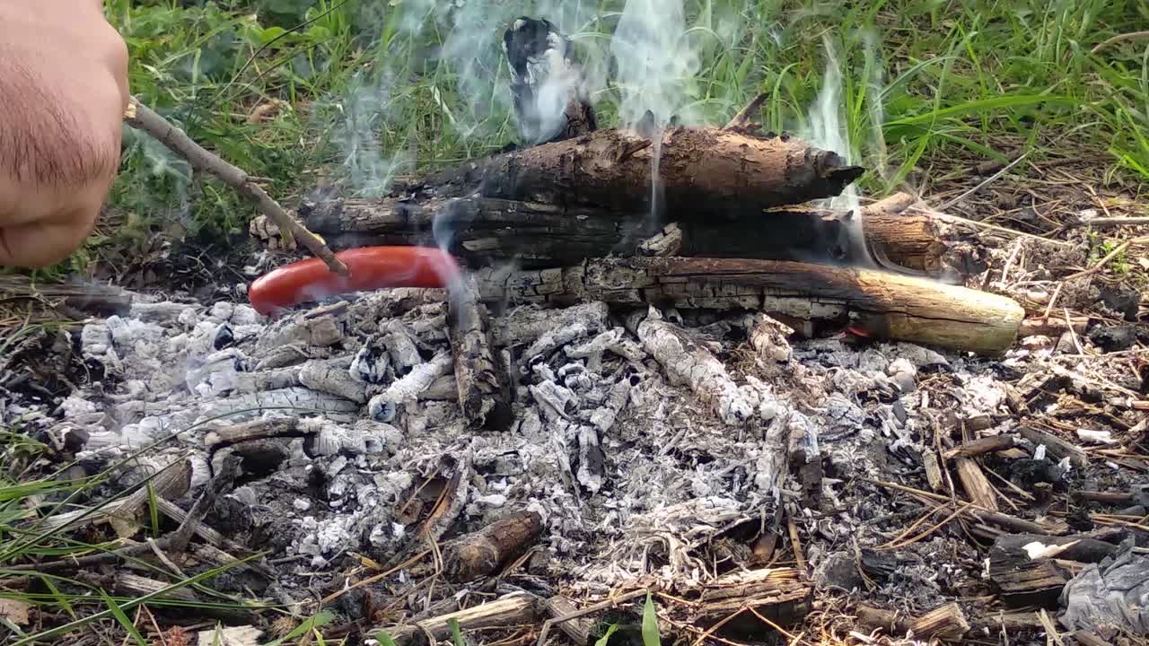Sausage roasting on fire while hiking the mountains at summer in a bright summer day. A man cooking snacks on small bonfire. POV view.