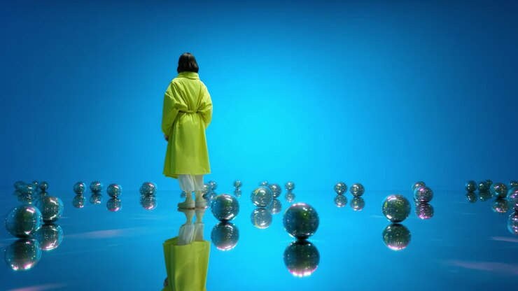 A solitary figure in a bright yellow coat stands contemplatively amidst a surreal landscape of reflective spheres, embodying themes of solitude, introspection, and vibrant imagination against a stunning blue backdrop