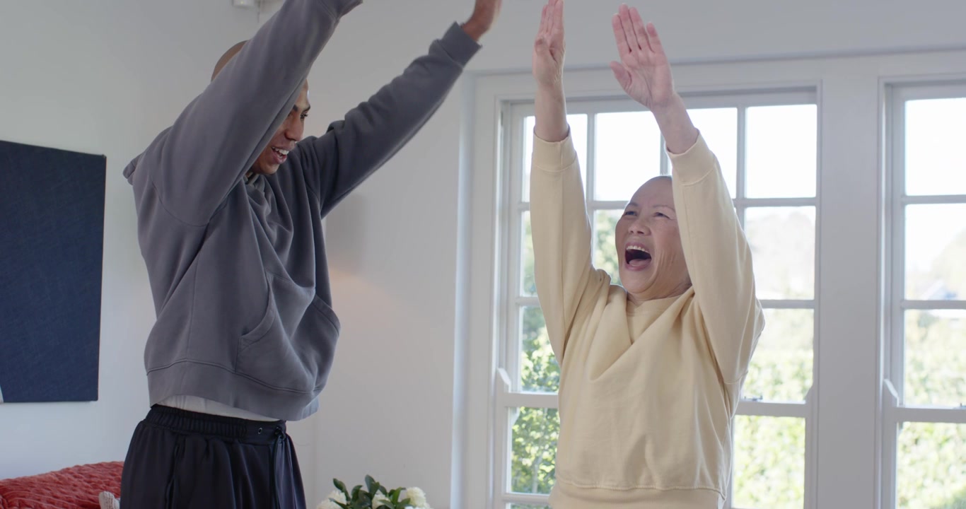 Senior Asian woman and African American man smiling and doing arm stretches by window for mobility
