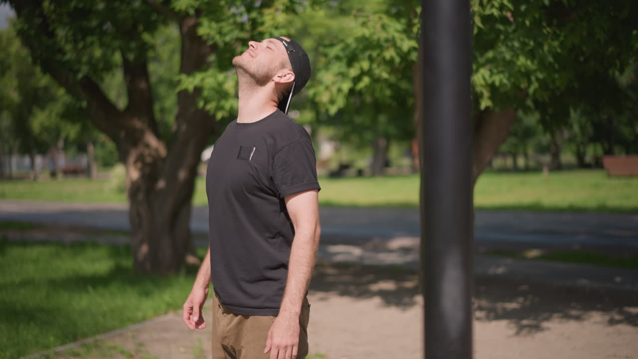 Man Engaging In Mobility Exercises, Person Slowly Stretching Neck And Shoulders Outdoors Before Workout, Man Carefully Executing Mobility Stretches Beside Bars In Leafy Park Environment