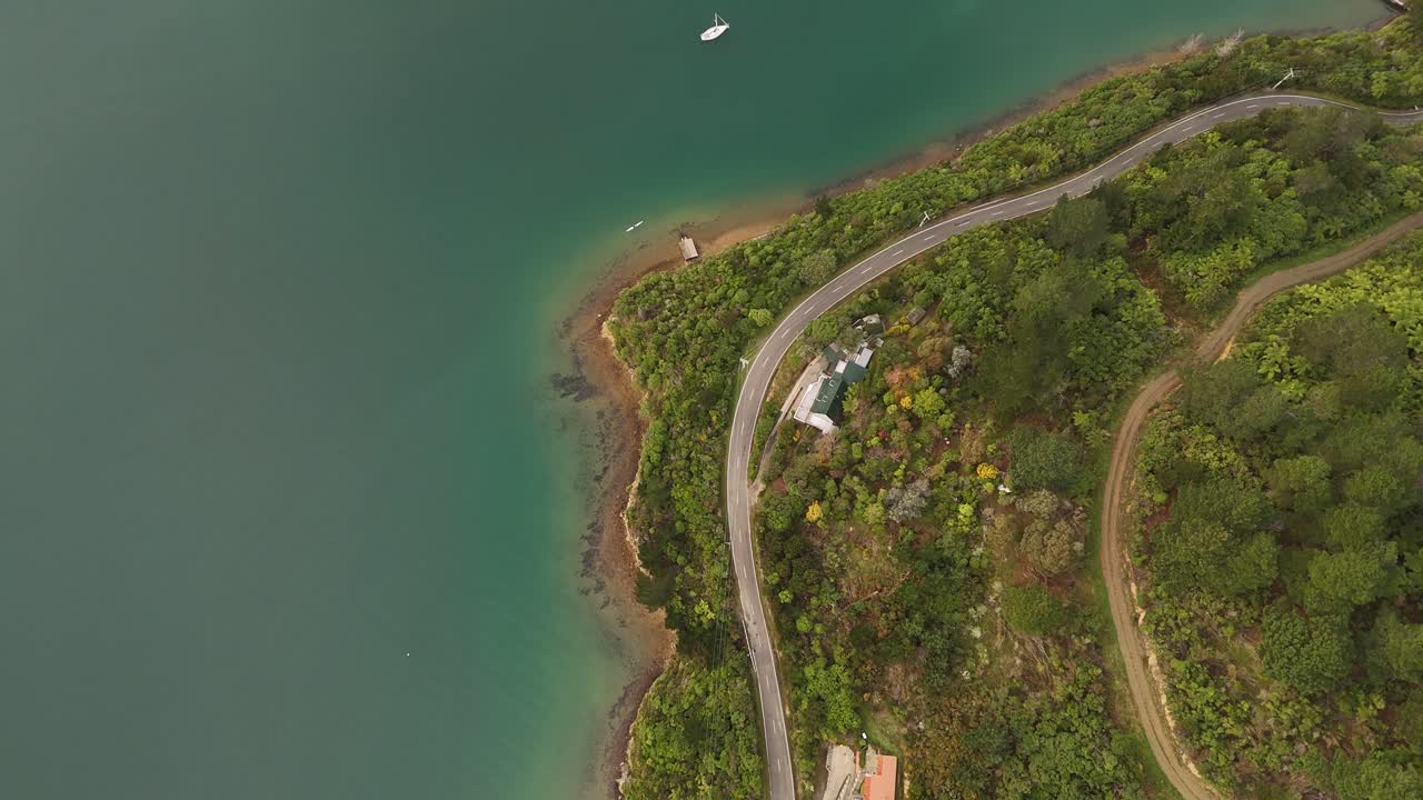 Scenic coastal road alongside calm water of Marlborough Sounds, house nestled lush bush, kayak near shore, South Island, New Zealand. Aerial top-down view