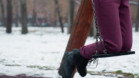 lower body view of person in maroon trousers and black boots gently swinging, surrounded by snow, blurred background with bare trees, building, and passing car