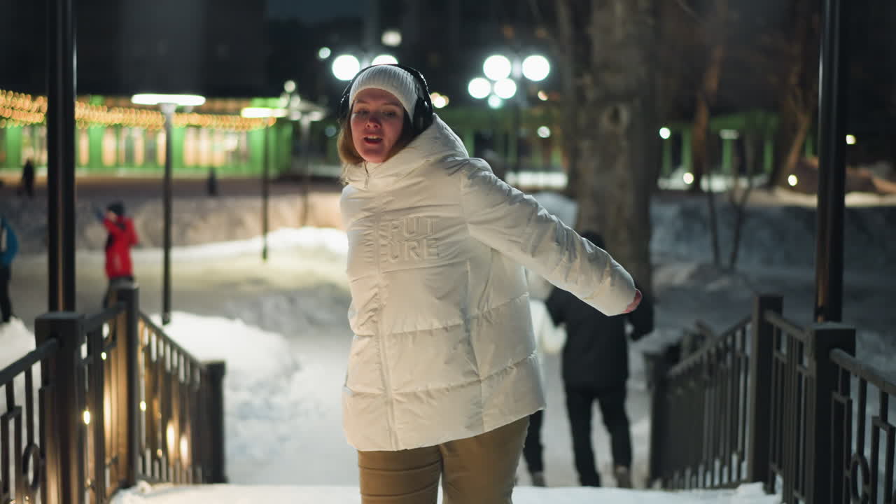 Close Up Of Girl Dancing On Pavilion Stairs Under Fairy Lights At Night, Twirling In White Puffer Coat And Headphones, Joyful Movement Across Fresh Snow In Illuminated Park Scene