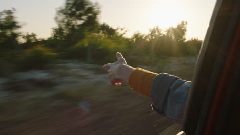 Frau-Im-Auto-Hält-Die-Hand-Aus-Dem-Fenster-Und-Spürt,-Wie-Der-Wind-Durch-Die-Finger-Weht,-Während-Sie-Im-Sommerurlaub-Auf-Dem-Land-Unterwegs-Ist-Und-Bei-Sonnenuntergang-Die-Freiheit-Auf-Der-Straße-Genießt