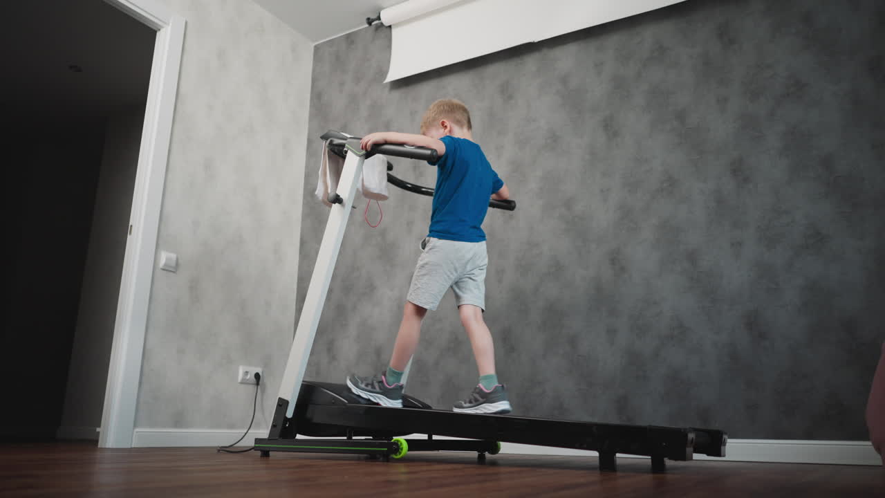 Back view of little child walking on treadmill holding handles during exercise session in home gym room with grey textured wall wooden floor power cord visible pink beanbag in corner