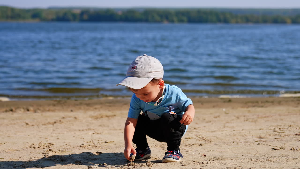 Cute baby boy sits at the beach looking for the stones. Child heads to the water to throw the pebbles.