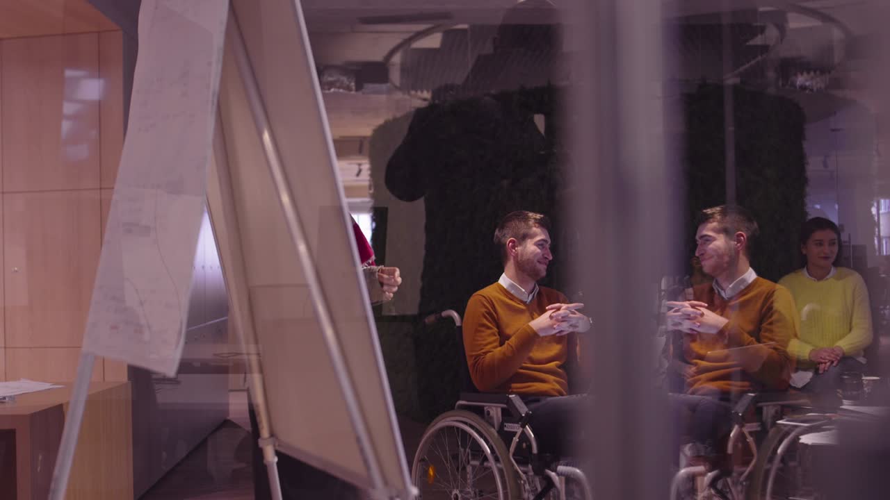 Muslim woman in a modern open space coworking office giving a presentation to team. Middle eastern Arab businesswoman in creative startup office. Disabled businessman in a wheelchair on team meeting