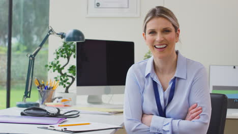 portrait of smiling female doctor or gp in office sitting at desk with folded arms