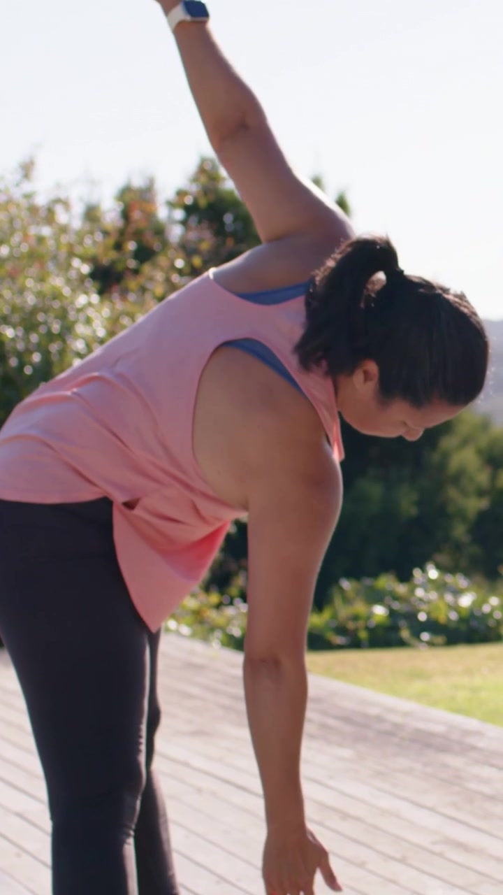 Video vertical: Mujer con top rosa, siguiendo una señal del guía, haciendo una flexión lateral en una terraza con una esterilla verde.