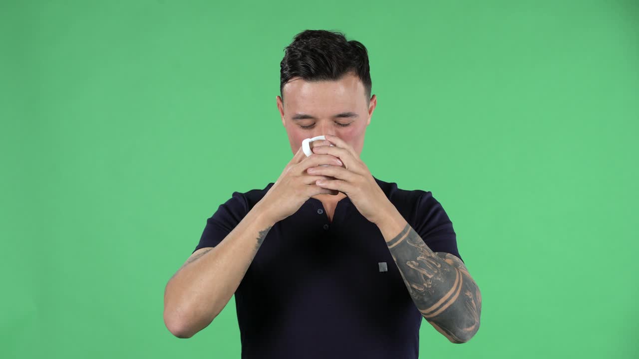 잘생긴 젊은 남자의 초상화는 직선적으로 보이고 커피를 즐기고 있습니다. 검은 티셔츠를 입고 손에 문신을 한 스타일리시한 머리카락을 가진 갈색을 태우고 스튜디오의 녹색 화면에