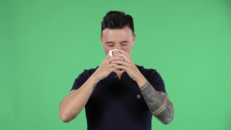 retrato de un joven guapo está mirando directo y disfrutando del café. morena ardiente con cabello elegante en una camiseta negra y tatuajes en las manos en una pantalla verde en un estudio