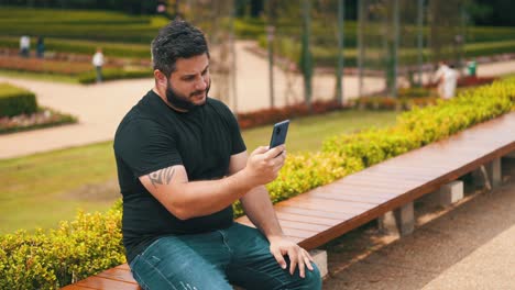 Young-bearded-man-on-jeans-pants-using-his-cell-phone-outdoors-while-seated-on-a-bench