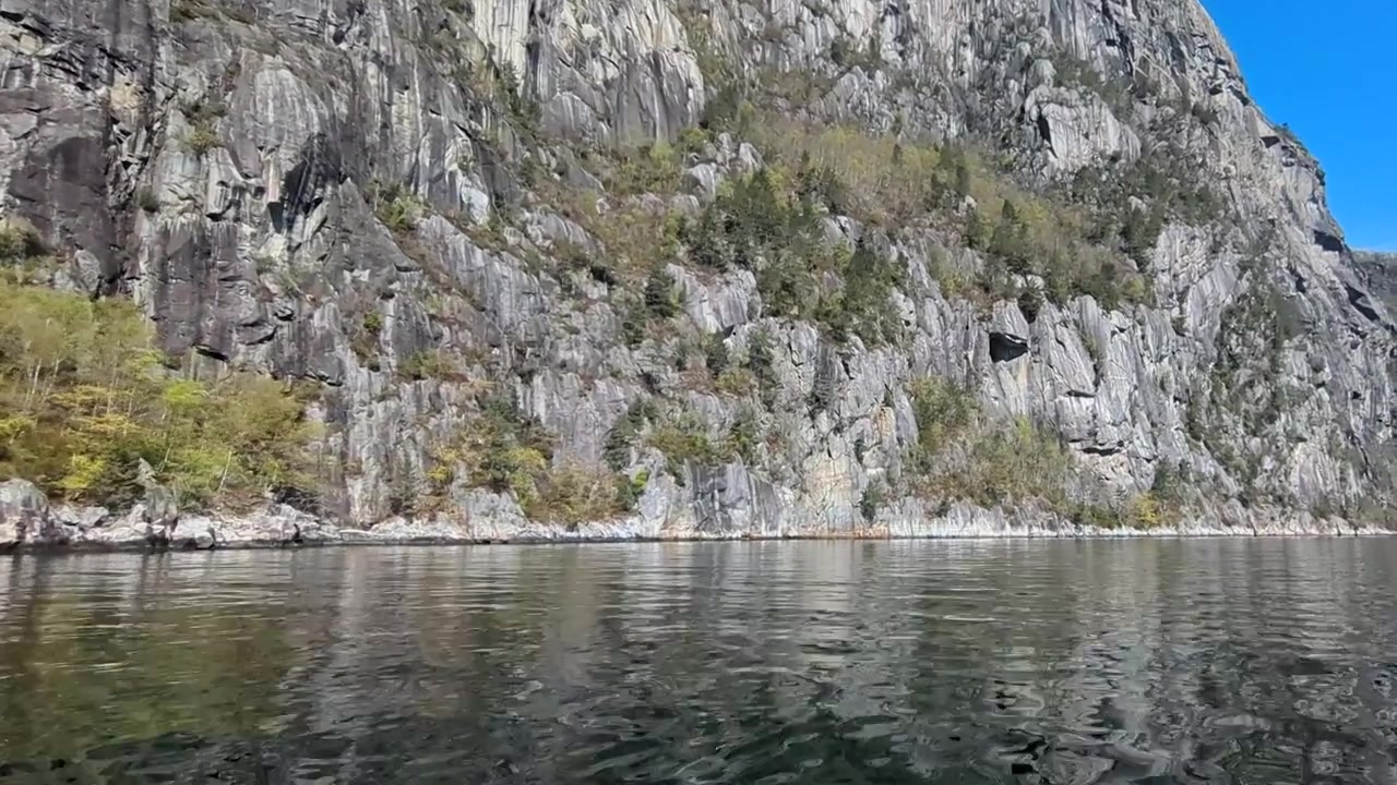 Steep Rocky Mountain Cliff Rising from Calm Blue Water with Distant Snow Capped Peaks Norway