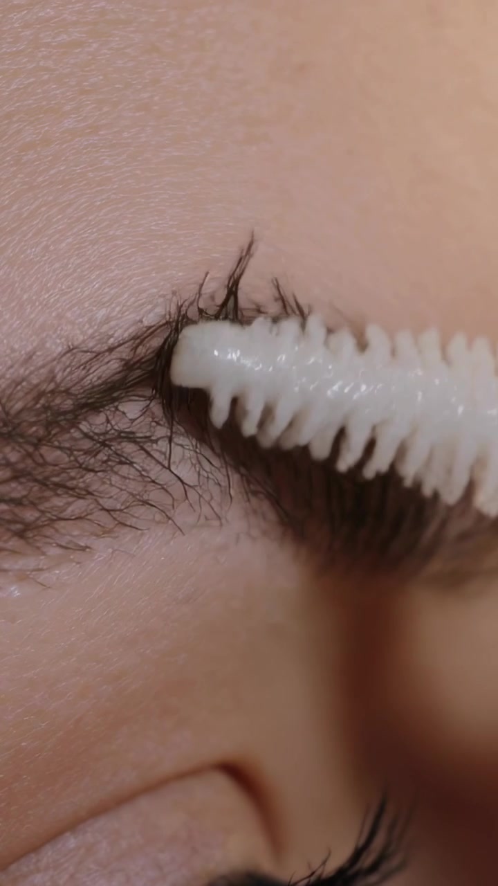 Vertical video: Spoolie approaching, brushing right eyebrow to shape hairs with white gel at vanity