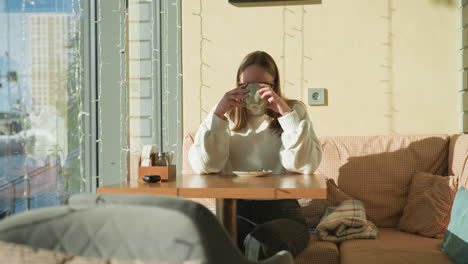 writer dressed in white sweater seated by cafe window sipping coffee with thoughtful expression, sunlight casting soft shadows on wall, glass reflections and urban view outside