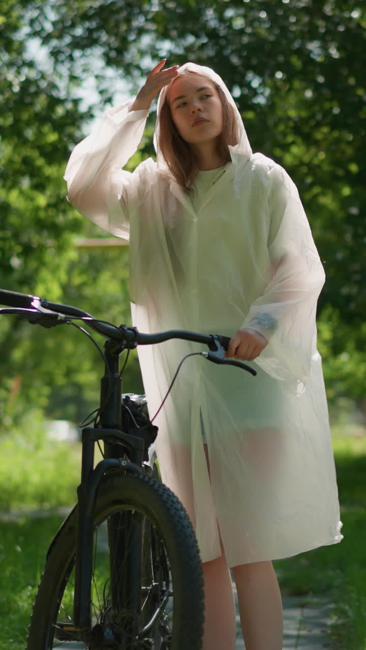반투명한 레인코트를 입은 젊은 여자가 자전거를 타고 빛의 길을 따라 멈추고, 기둥 근처에 스탠드를 놓고 후드를 제거하여 바닥에 사려 깊게 기대고, 에 손을 고, 초록색 배경으로