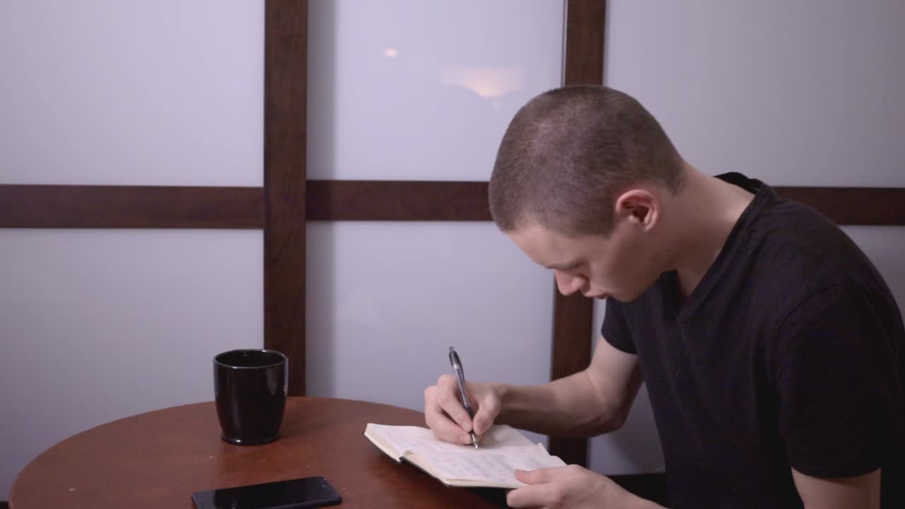 Man Wearing A Casual Black Shirt - Writing With Pen On Notebook In The Coffee Table