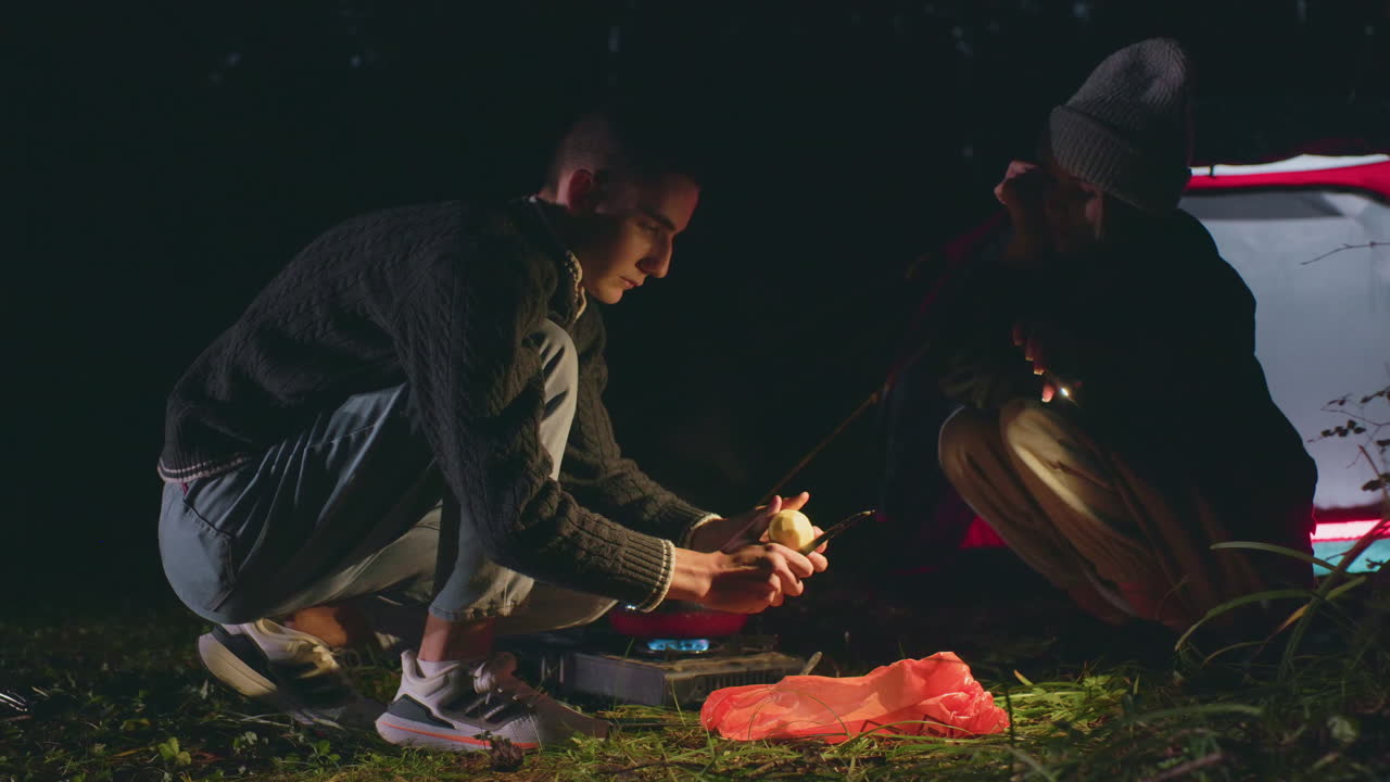 Woman uses flashlight to light up area while man peels potato during night camping in forest, both crouched near tent and portable stove, focused on food preparation
