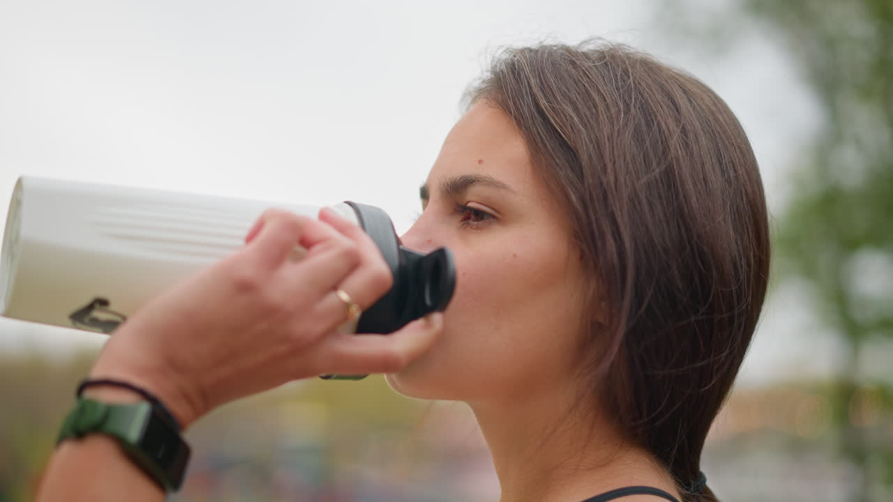 Close-up shot of lady drinking water from white bottle with wrist watch and ring on her hand, showcasing hydration and wellness during outdoor exercise or workout