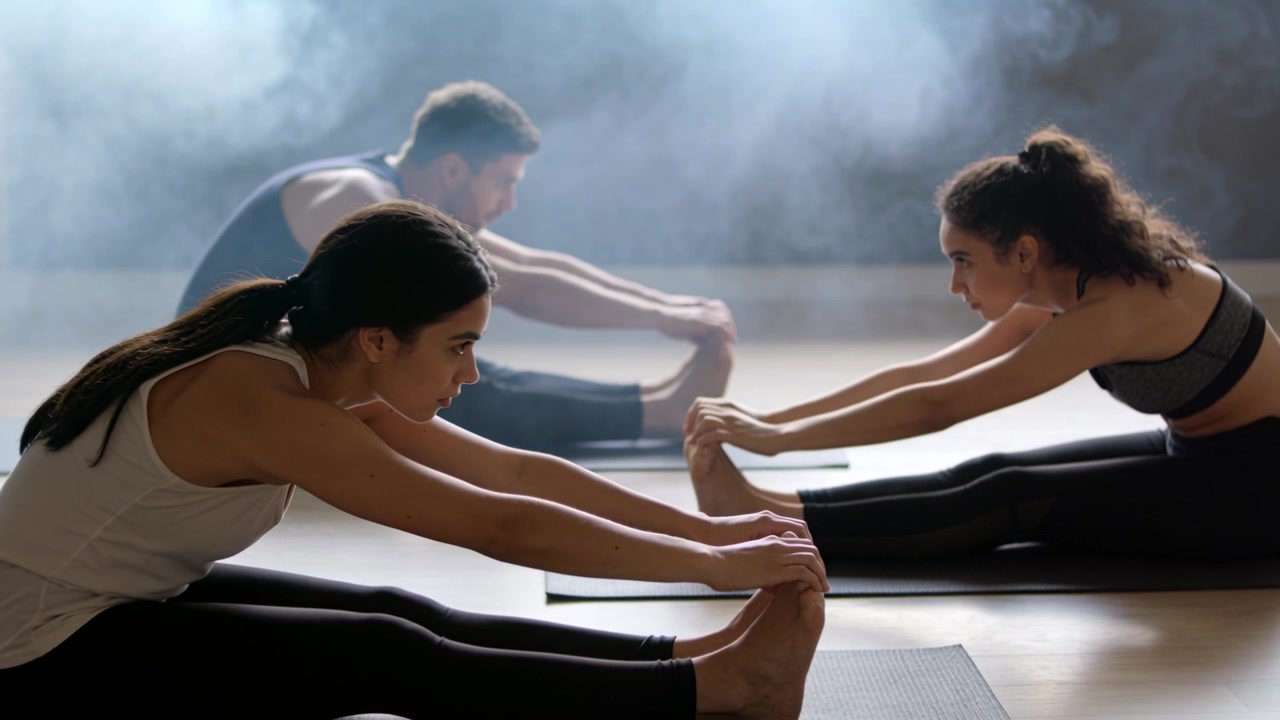 Leaning three adults in workout wear deepening seated forward-fold on mats in studio, phone nearby