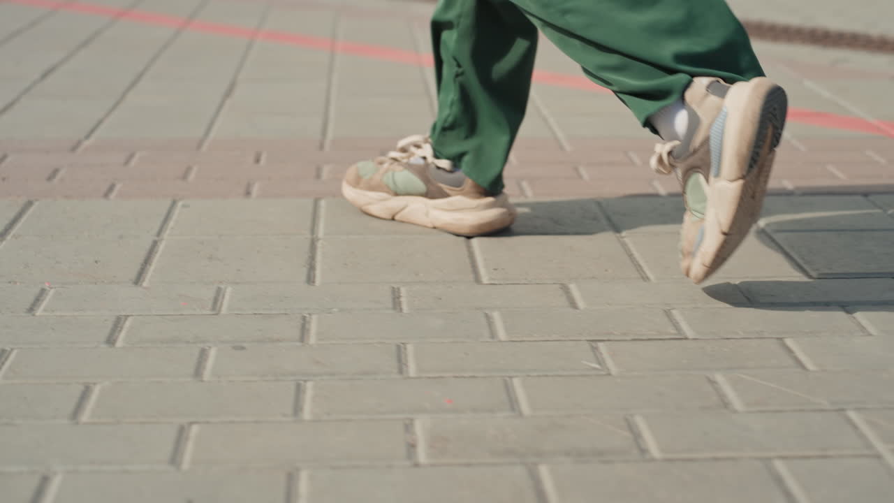 closeup feet on pavement green trousers steady determined stride across textured sidewalk tiles, chunky sneakers scuffing ground, soft morning light and long shadow lines, urban routine, suggested