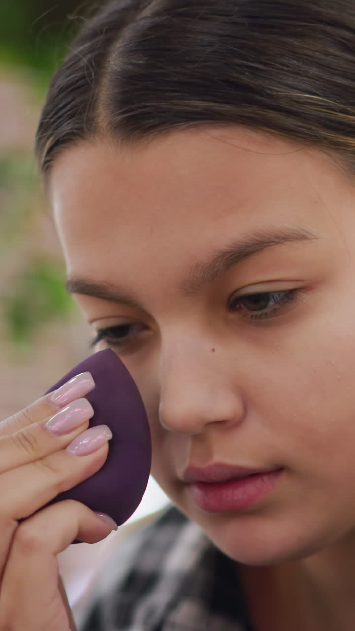 primer plano y tomas de tres cuartos de una adolescente difuminando maquillaje con una esponja en un jardín iluminado por el sol, mostrando una práctica concentrada de contorno e iluminación, movimientos suaves de presión, rosa