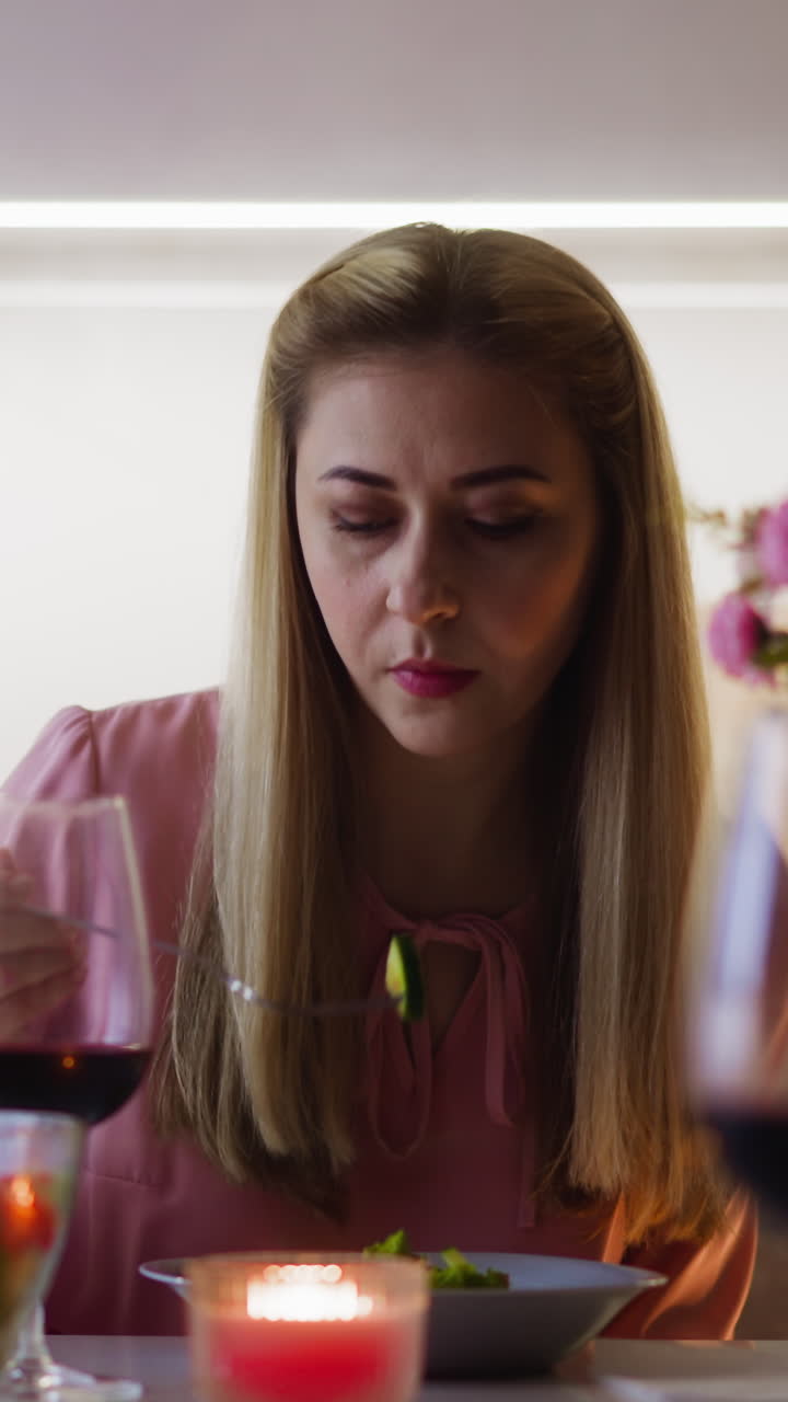 una mujer rubia bonita en un vestido elegante con una sabrosa copa de vino rojo come una ensalada fresca y jugosa sentada en la mesa en una cena festiva en la cocina en cámara lenta