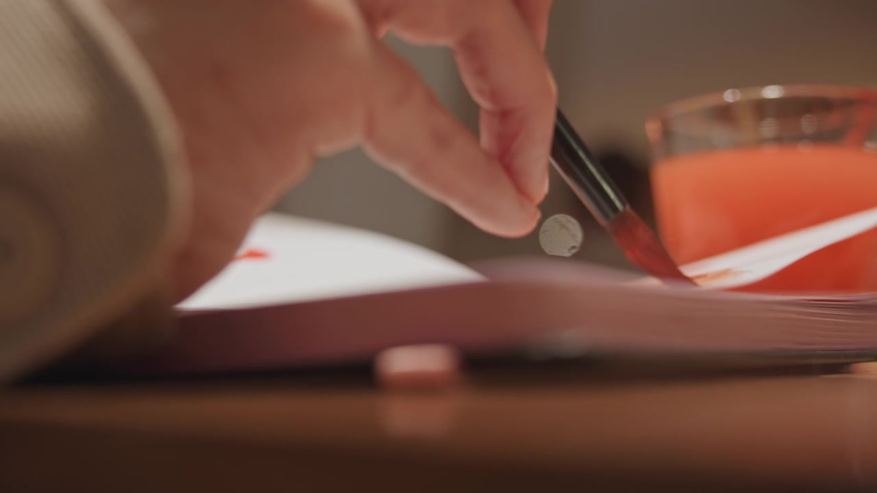 Close up of hand holding paintbrush applying red liquid to paper surface. Orange glass with pinkish orange contents visible in background on wooden table. Soft lighting creates warm ambiance