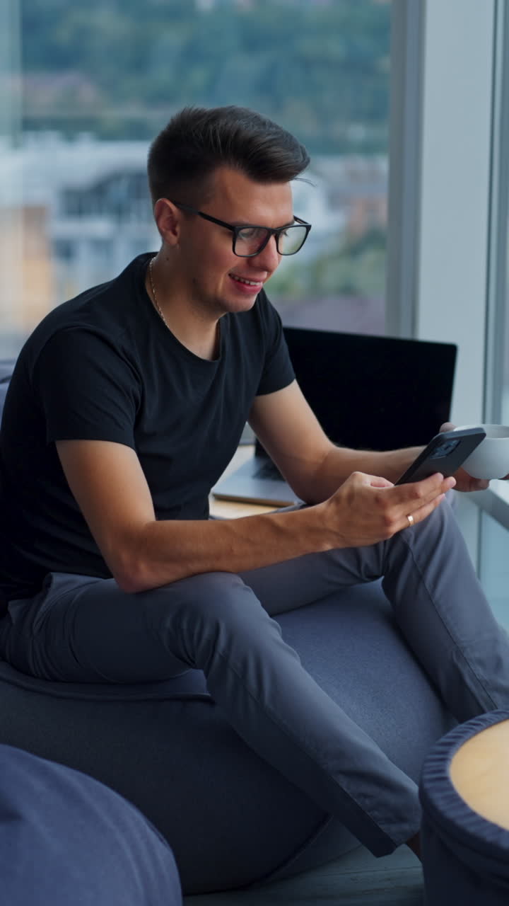 Dark-haired man in glasses sitting on bean bag chair holding a phone and a cup. A break from work at computer. Panoramic windows in blur at backdrop. Vertical video