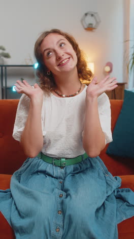 happy young woman vlogger smiling friendly at camera and waving hands while sitting on sofa at home