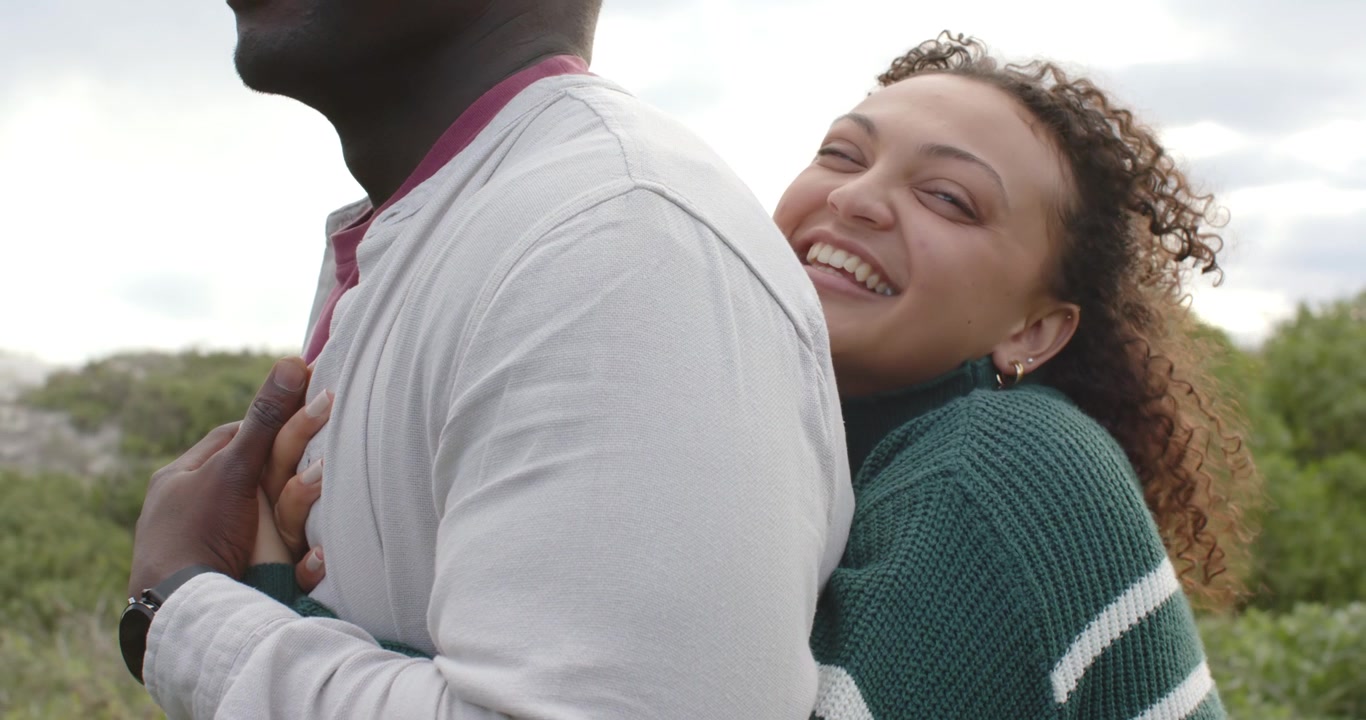 African American couple hugging in field, man placing hand over her hands, making her laugh, watch