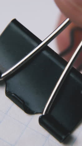 person fixes black metal clamp on checkered paper sheets on white background extreme close view