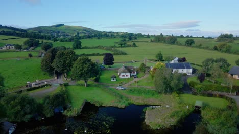 Pintoresca-Hierba-Verde-Colorida-Y-árboles-En-El-Campo-Irlandés-Al-Lado-De-Un-Lago-Con-Una-Cabaña-Tradicional-En-La-Línea-De-La-Costa,-Drones-Sobrevuelan-En-Un-Día-Soleado-Y-Brillante