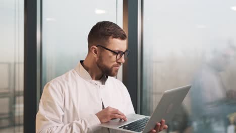 Portrait-of-a-male-doctor-in-a-white-lab-coat-with-a-laptop-in-his-hands-standing-by-panoramic-windows-in-a-modern-new-clinic