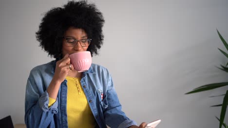 Mujer-Negra-Riendo-Usando-Un-Teléfono-Inteligente-Y-Bebiendo-Una-Taza-De-Café