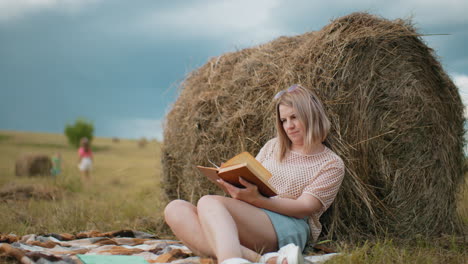 joven con gafas de sol en la cabeza descansando contra la bala de heno, leyendo un libro en el tranquilo campo, vista borrosa de los árboles y los niños jugando en la distancia