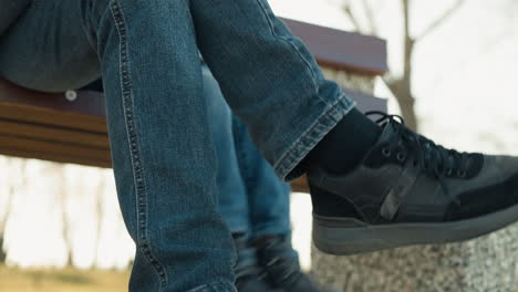 close-up of an adult and a child sitting closely together on a bench, with the child gently hitting the adult's crossed leg, both are wearing jeans and boots, with the adult have a black socks