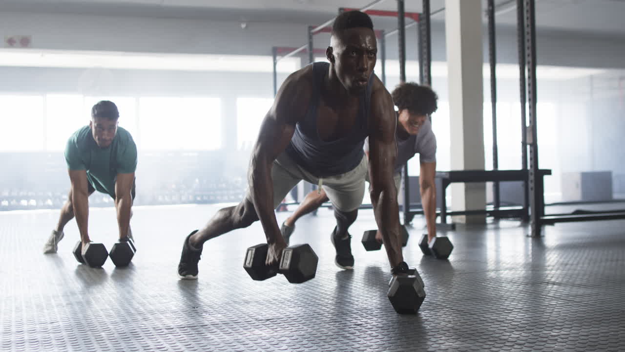 Diverse young men training with dumbbells in gym, doing push-up exercises