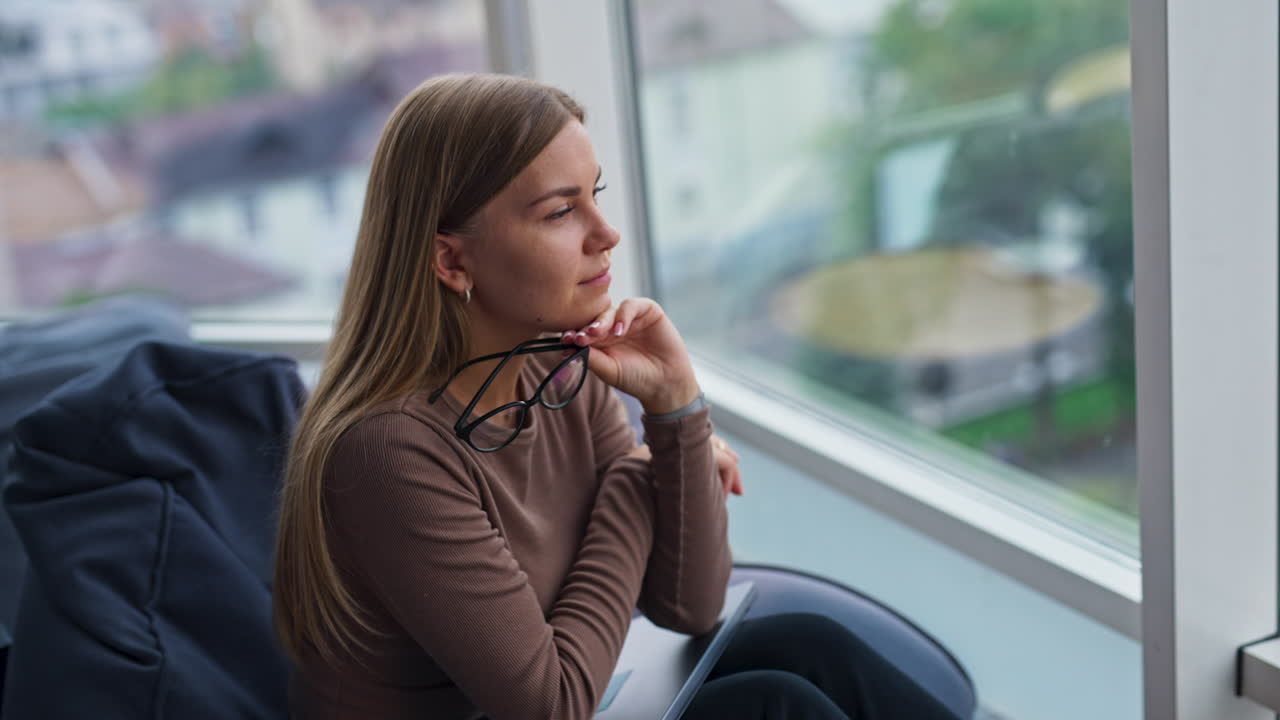 Tired thoughtful blonde lady resting from work on laptop. Young lady puts on her glasses and smiles to the camera. Blurred backdrop.