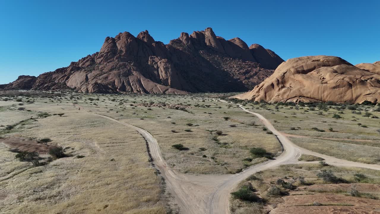 Drone aerial view of dusty desert tracks winding through arid plains towards the granite peaks of Spitzkoppe in Namibia, under a deep blue clear sky