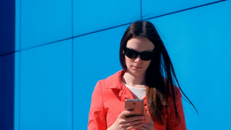 young woman brunette in sunglasses in red coat waits for someone and checks her phone, texting.