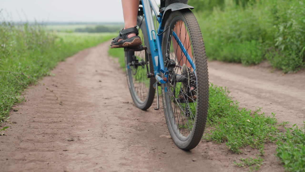 chico blanco en bicicleta con gran angular en una carretera rural, cadencia constante sobre grava blanda, huellas de neumáticos que conducen a un horizonte distante, setos exuberantes y campos abiertos, ritmo exploratorio relajado con luz diurna ambiental