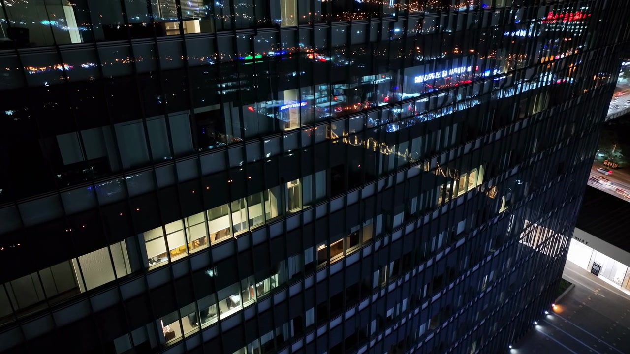 Vista noturna que se move constantemente para cima através de um exterior de vidro escuro, onde janelas de escritórios brilhantes e luzes coloridas da cidade se refletem em uma grade de painéis brilhantes e linhas estruturais.