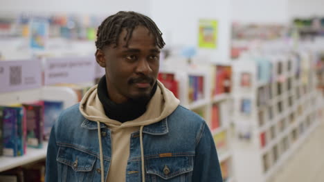 close-up of man smiling and looking around in store, dressed in denim jacket, hoodie, and scarf, with books and shelves visible in background, bright indoor lighting, and vibrant atmosphere