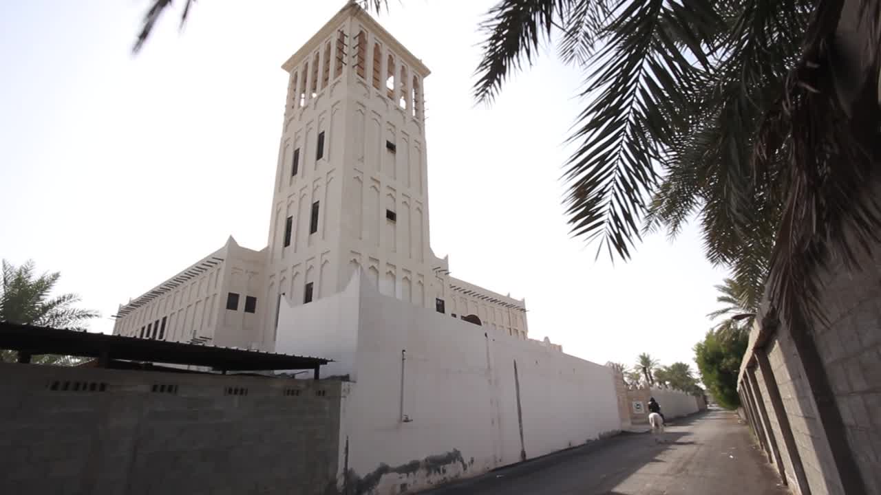 Slow parallax shot of an old Arabian tower with palm trees to the sides and the sun shines through the windows of the tower, in addition to a person riding a white Arabian horse seen from a distance
