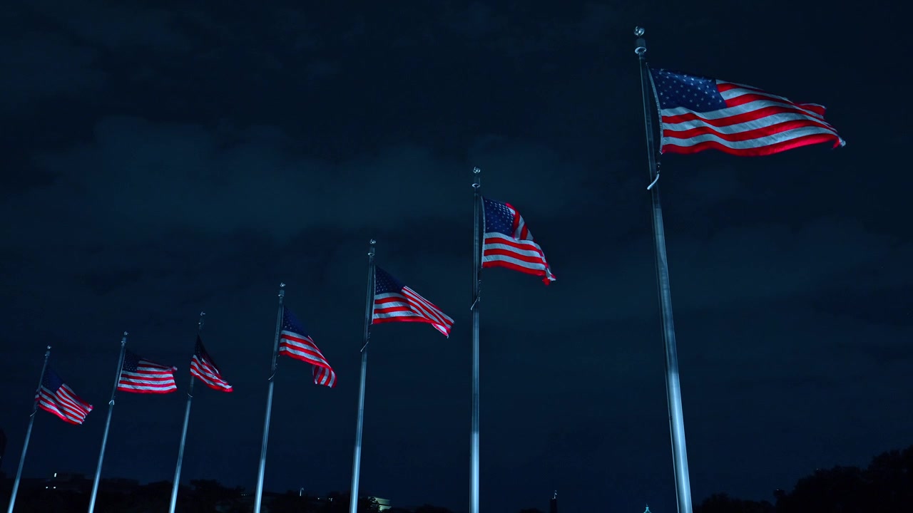 Row of American flags on flagpoles against dark night sky, Washington DC. Low angle view of flags waving in wind with cloudy overcast backdrop.