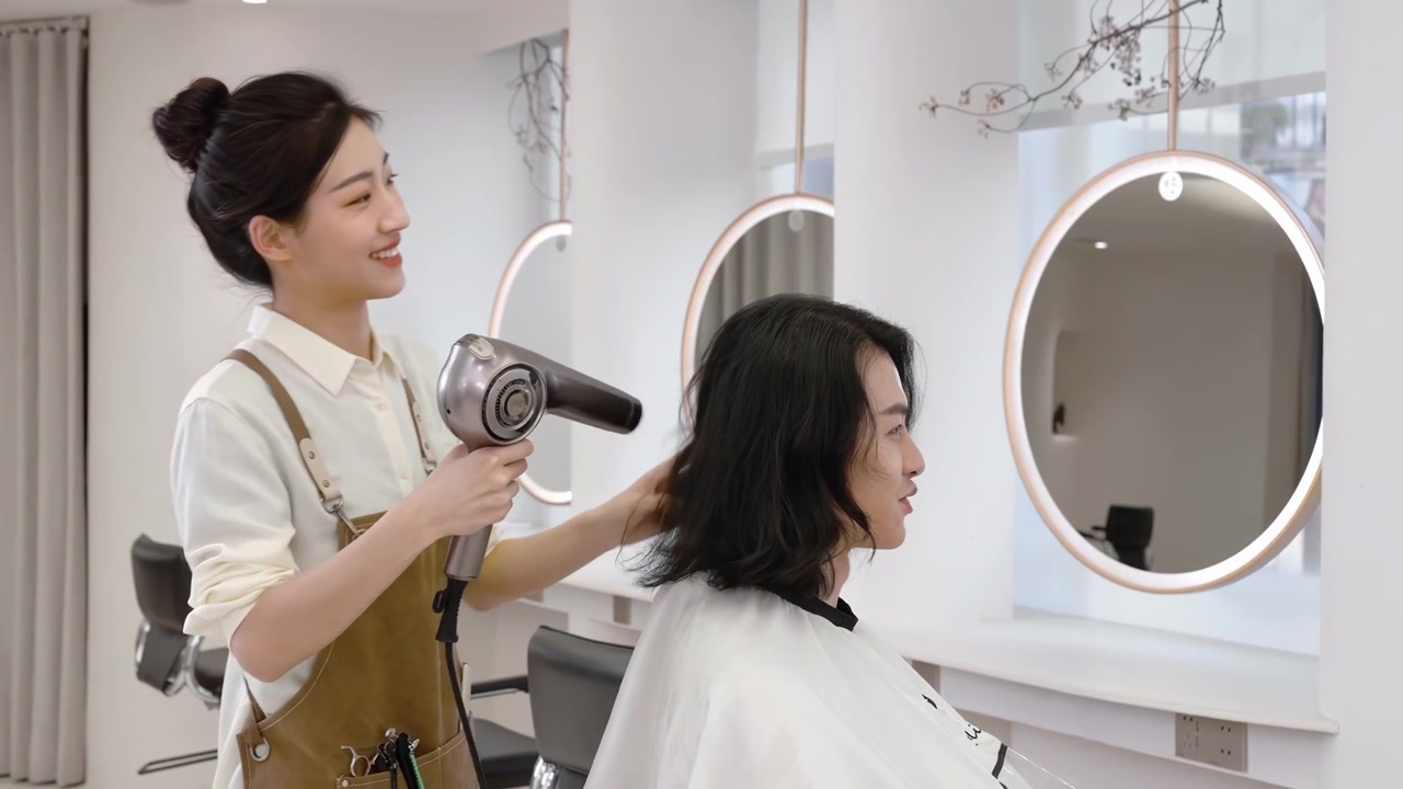 Parrucchiere stilista che asciuga e acconcia i capelli scuri di un uomo, utilizzando una spazzola rotonda e un asciugacapelli per lo styling; taglio di capelli e trattamento di bellezza per i clienti, con un mantello protettivo drappeggiato sulle spalle.