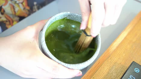 Toma-Lenta-De-Las-Manos-De-Una-Joven-Batiendo-Y-Mezclando-Té-Verde-Matcha-En-El-Tazón-De-Cerámica-Hecho-A-Mano-Con-Batidor-De-Bambú,-Matcha-Ceremonial-Tradicional-Japonés,-Bebidas-Especiales