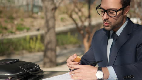 young bearded businessman eating snacks and writing notes, working with papers at park during lunch break