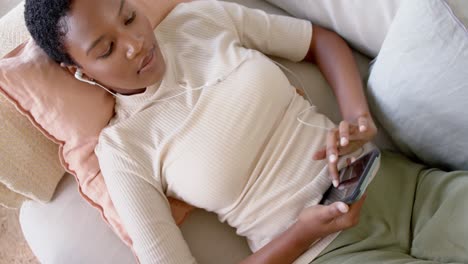happy african american woman lying on sofa wearing earphones and using smartphone, slow motion